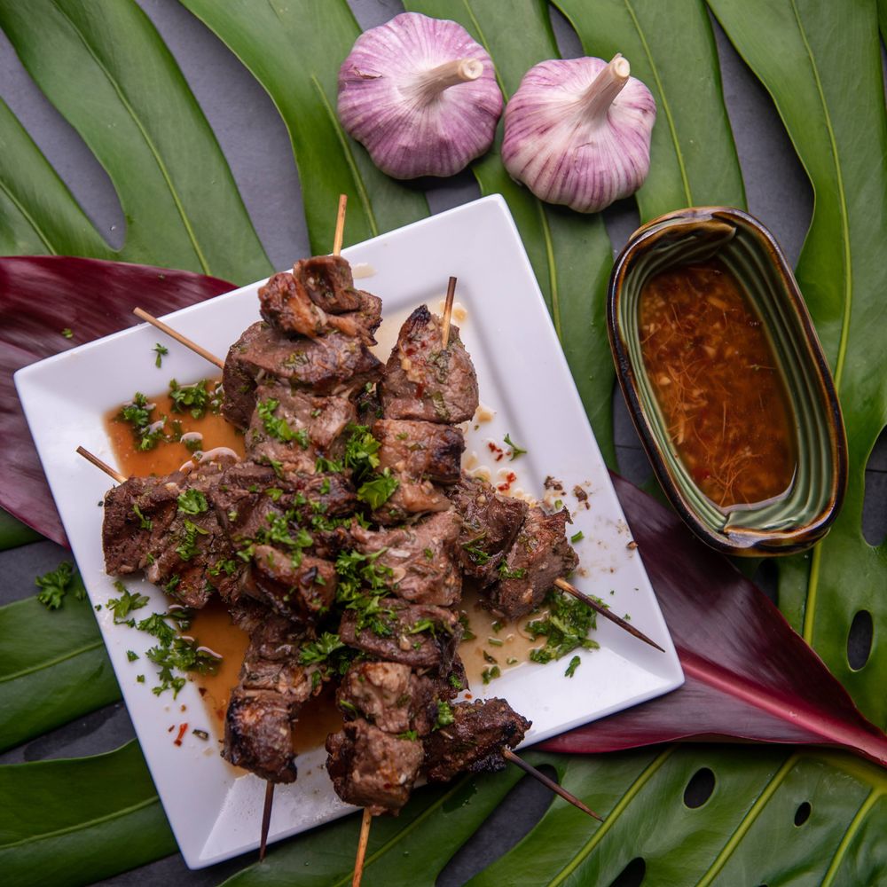 Grilled meat skewers with herbs and dipping sauce served on banana leaves with pink garlic bulbs.