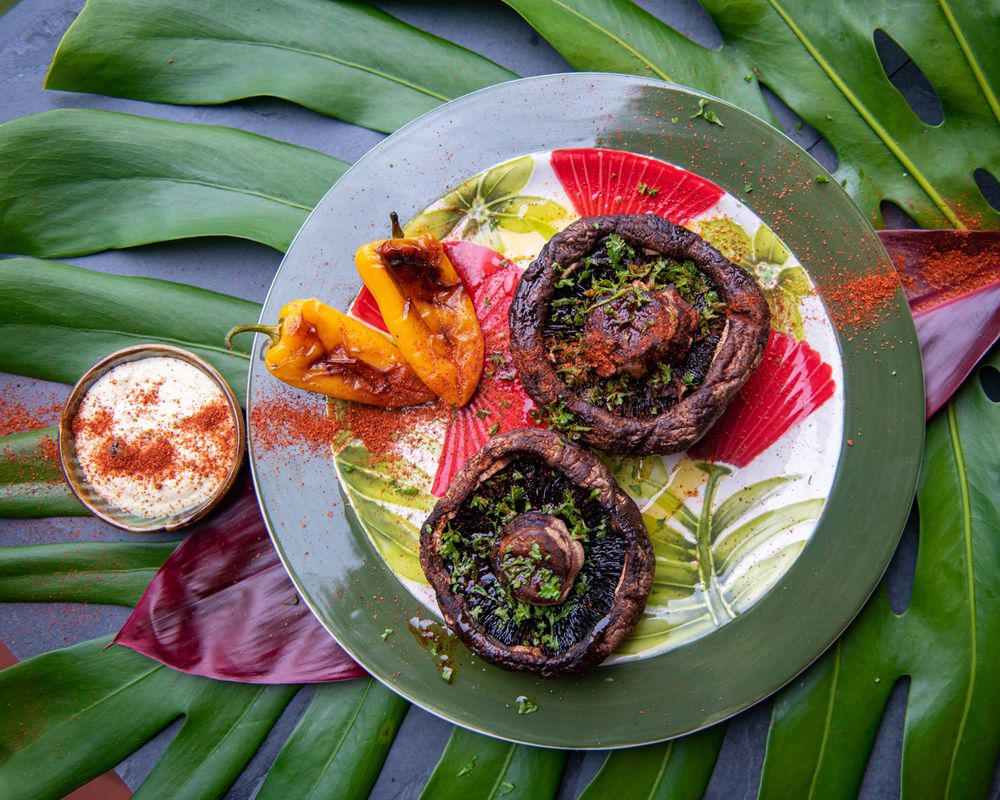 Grilled stuffed mushrooms with colorful garnishes served on a plate with tropical leaves and dipping sauce.
