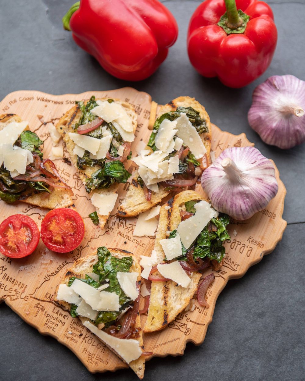Bruschetta topped with shaved parmesan and herbs served on wooden board with red peppers and purple garlic.