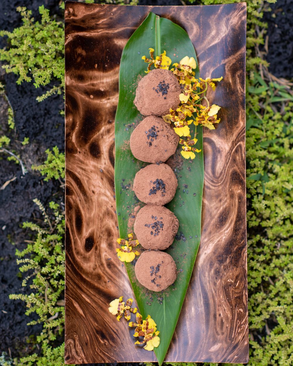 Chocolate truffles arranged on a banana leaf with yellow flowers and marbled background.