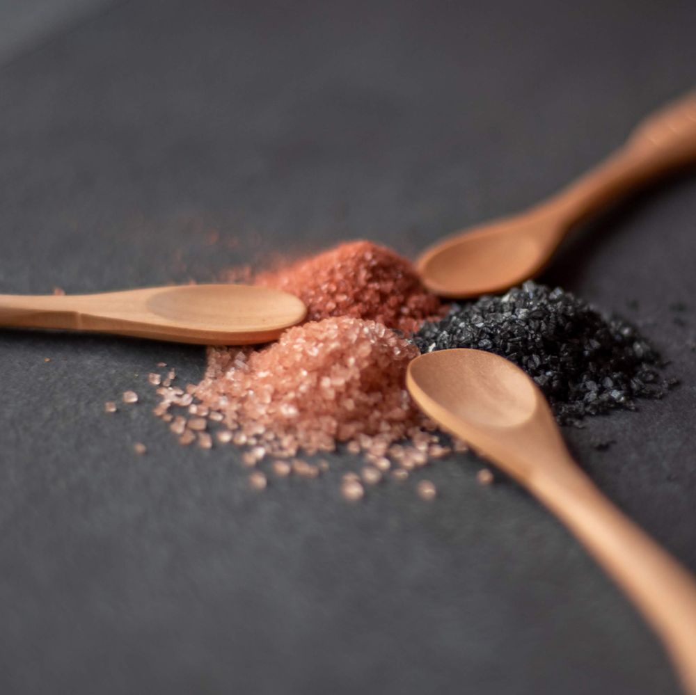 Three wooden spoons with pink Himalayan, black, and white sea salt on dark surface.