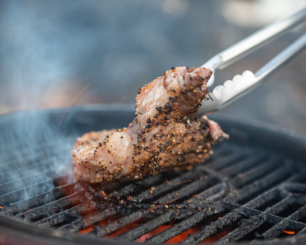 Grilled meat being lifted with tongs from a hot barbecue grill with smoke.