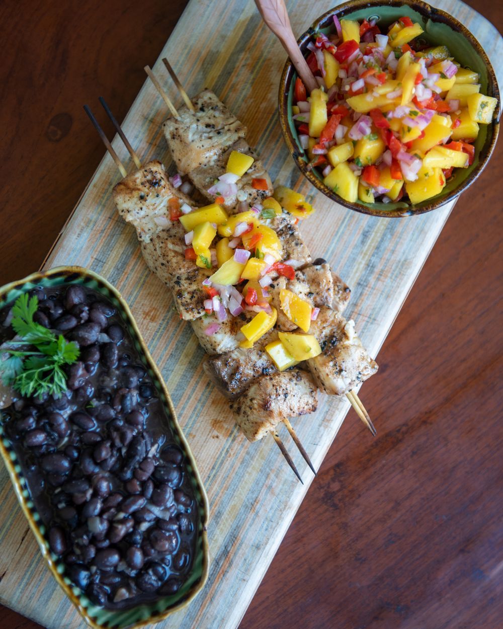 Grilled meat skewers with mango salsa and black beans served on a wooden board.