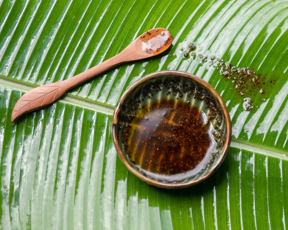 Dark liquid in coconut shell bowl with wooden spoon on green palm leaf.