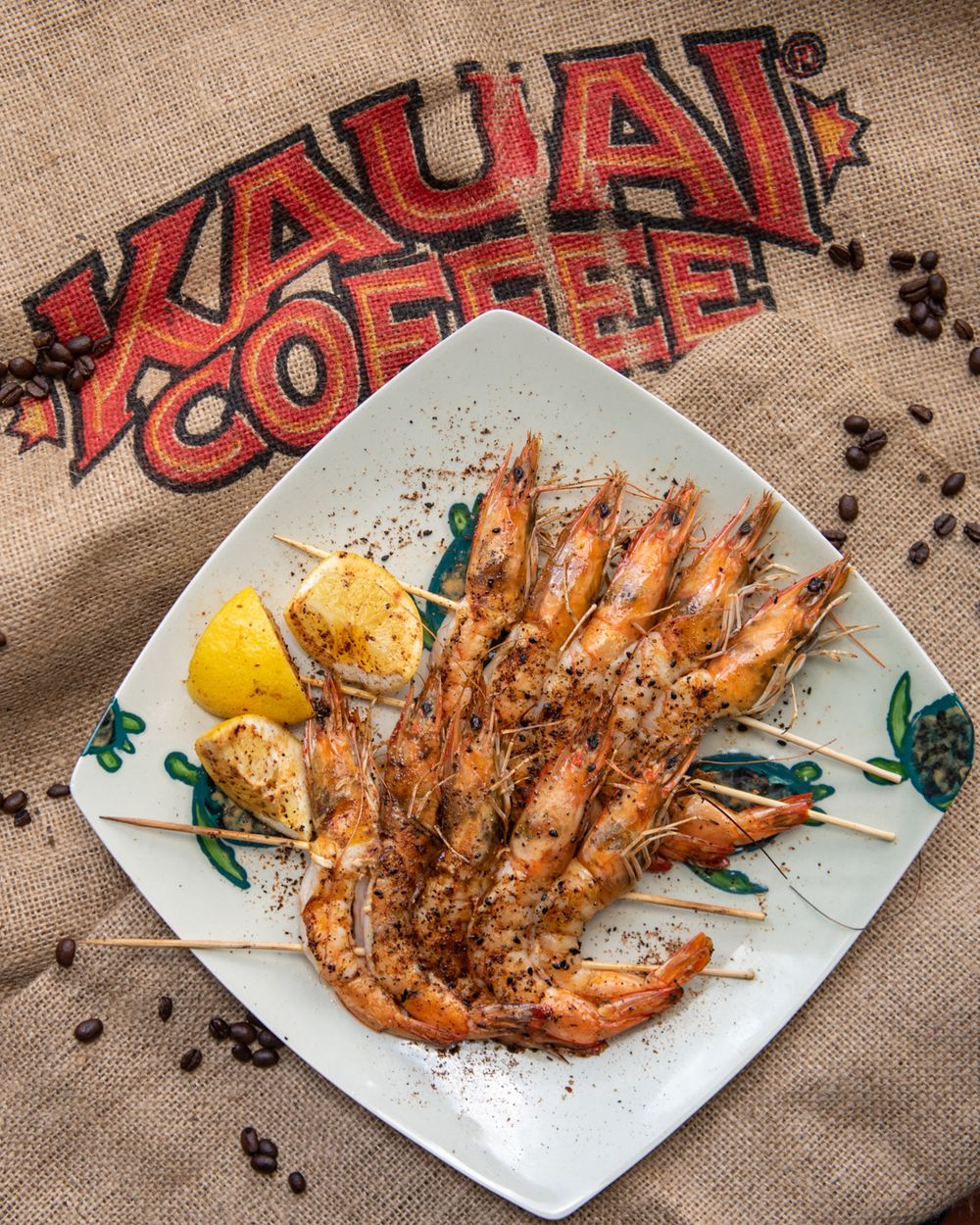 Grilled shrimp skewers with lemon on white plate against Kauai Coffee burlap background.