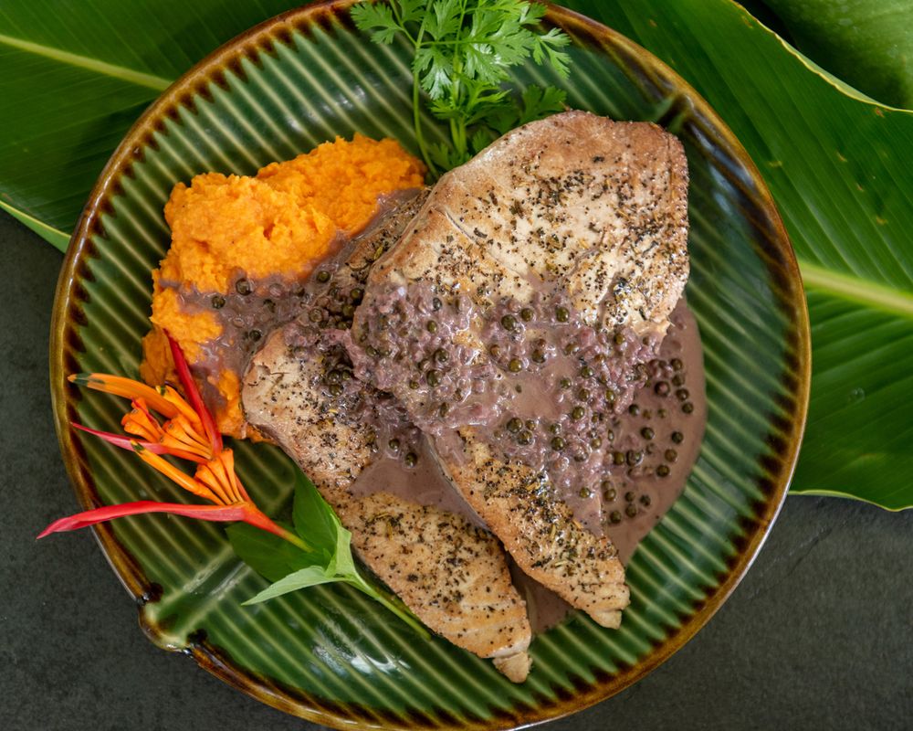 Seasoned fish fillet with orange puree and tropical garnish on striped plate with palm leaf backdrop.