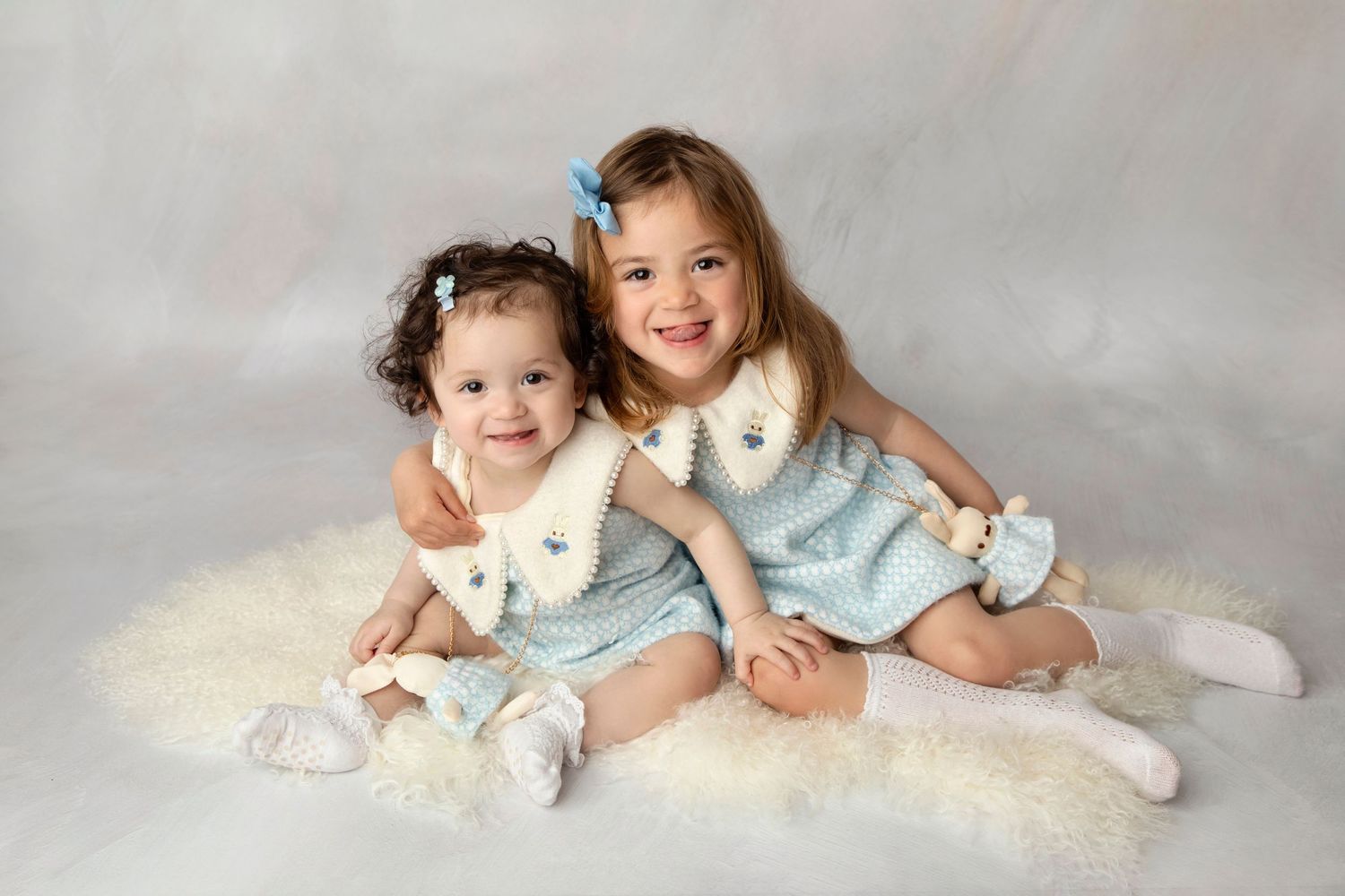 Two toddlers in matching blue dresses pose together on soft white backdrop.