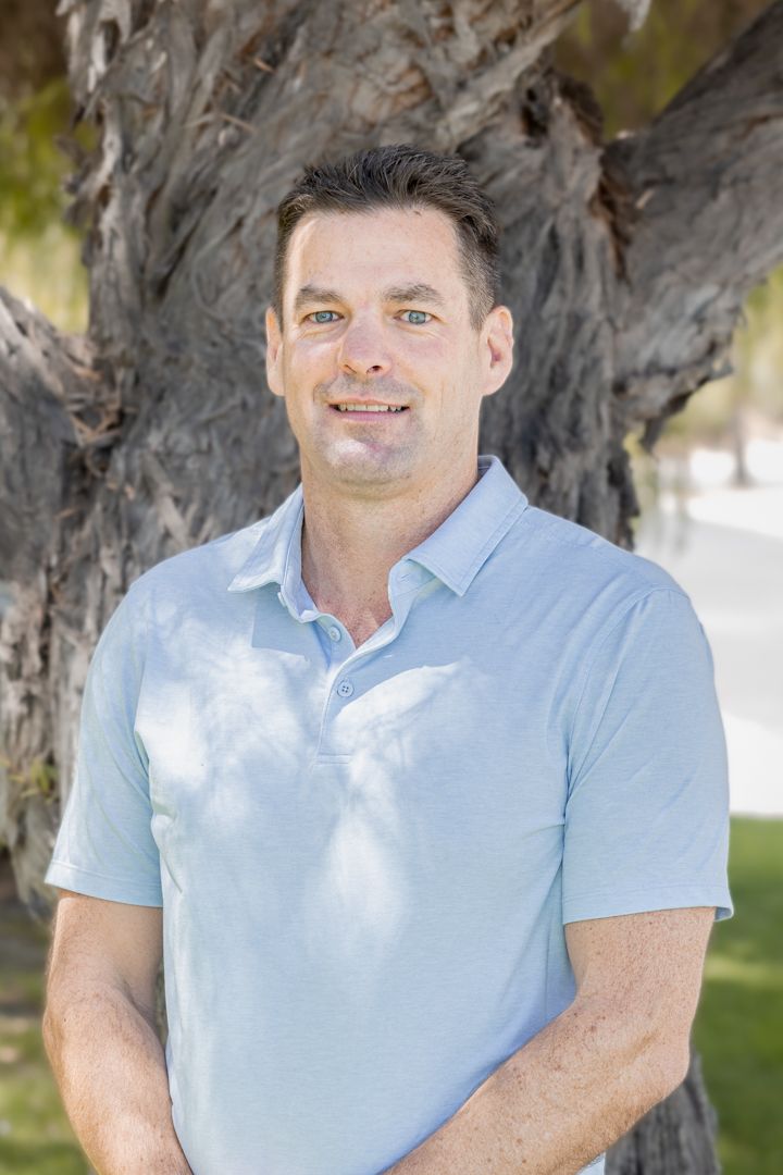 Professional headshot of someone in a light blue polo shirt standing in front of a large tree trunk.