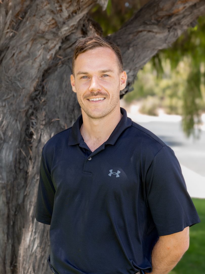 Athletic person in a navy Under Armour polo shirt smiles while standing next to a large tree.