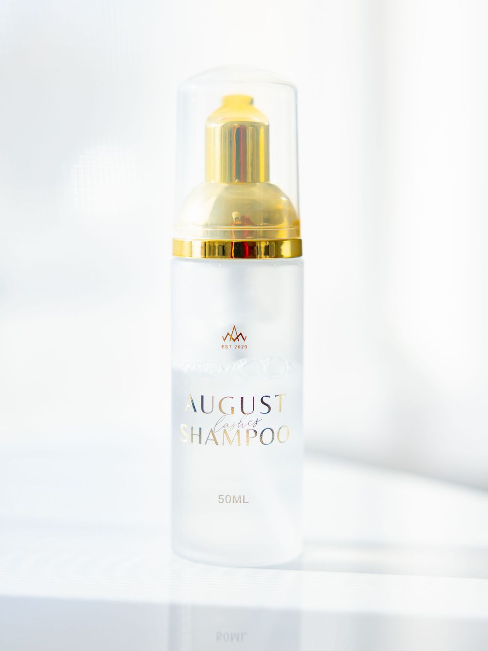 Elegant white cosmetic bottle with gold cap and 'August' branding against white background.