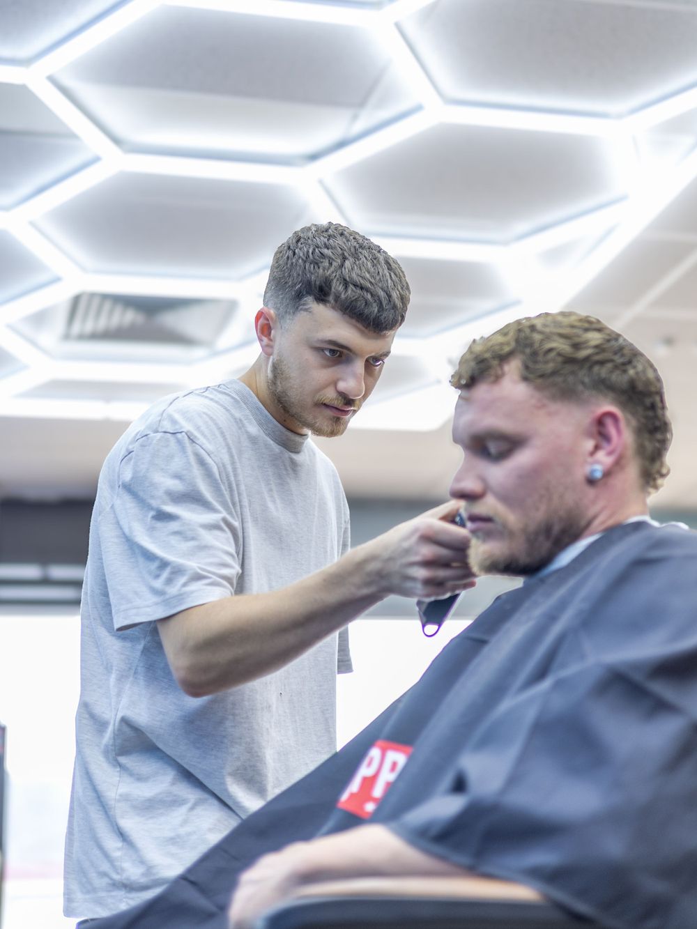 Professional haircut being given under modern hexagonal ceiling lights in a barbershop setting.