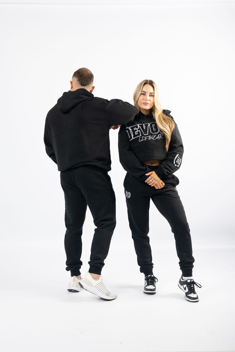 Two people wearing matching black hoodies and sweatpants posing against white background with athletic sneakers.
