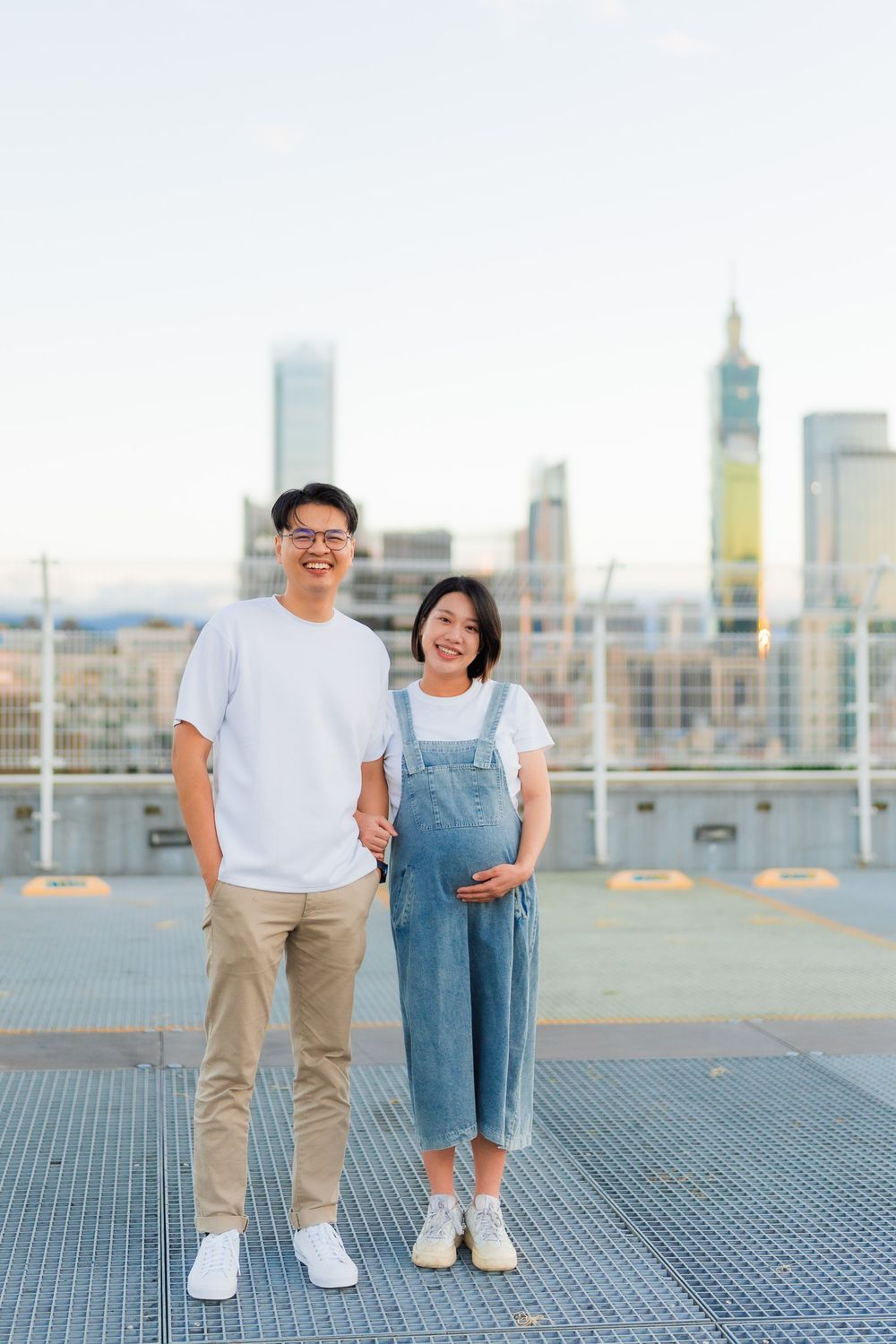Couple in casual attire standing on rooftop against city skyline backdrop at sunset.