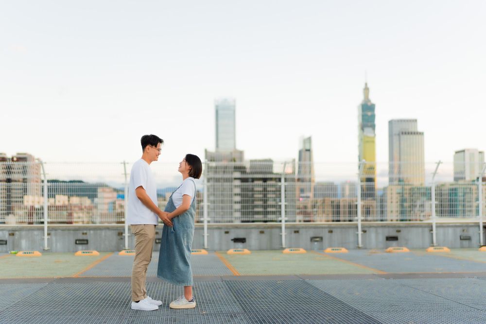 Couple standing together on rooftop with modern cityscape in background.