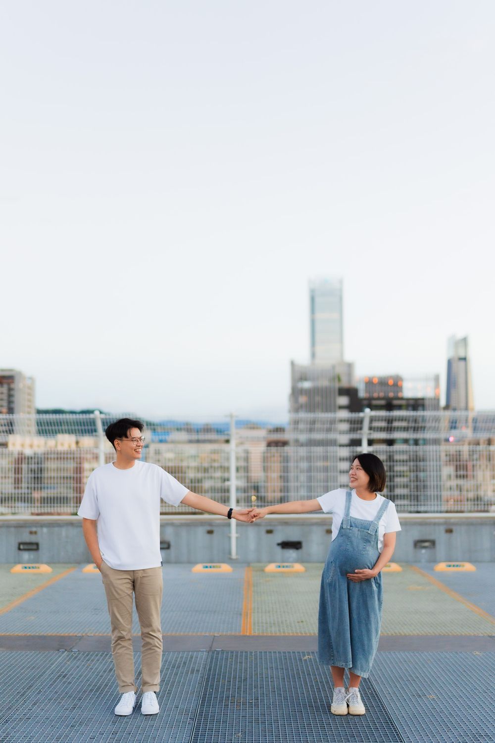 Couple stands apart on rooftop with cityscape background during maternity photoshoot in urban setting.
