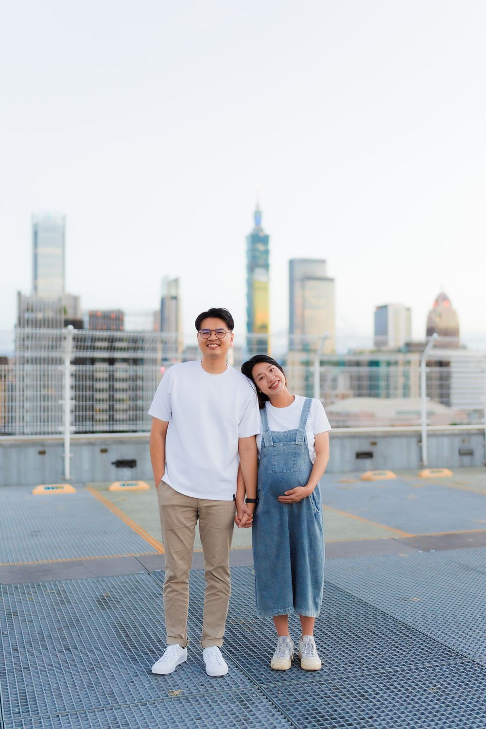 Couple poses together on rooftop parking deck with city skyline behind them at dusk.
