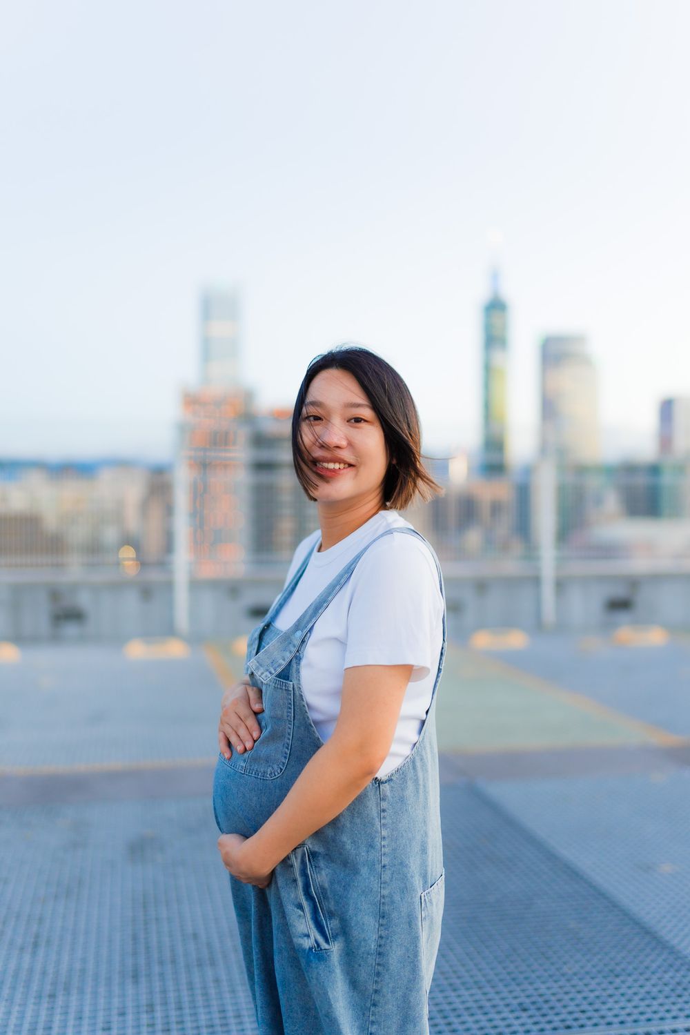 Expectant parent in denim overalls smiles while posing on rooftop with city skyline backdrop.