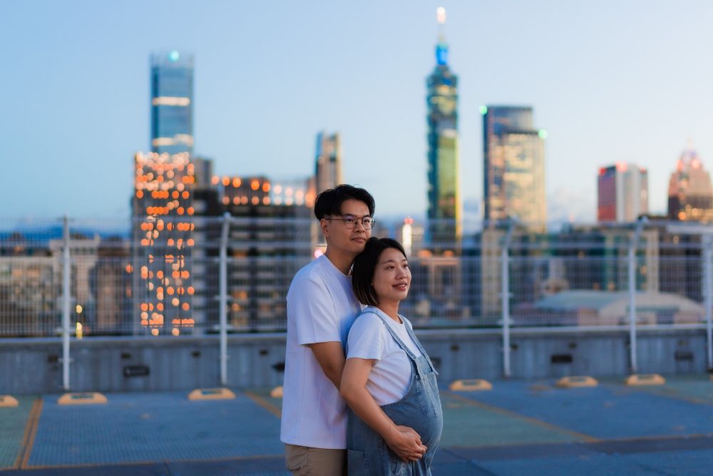 Couple shares tender moment during twilight maternity shoot with illuminated city backdrop.