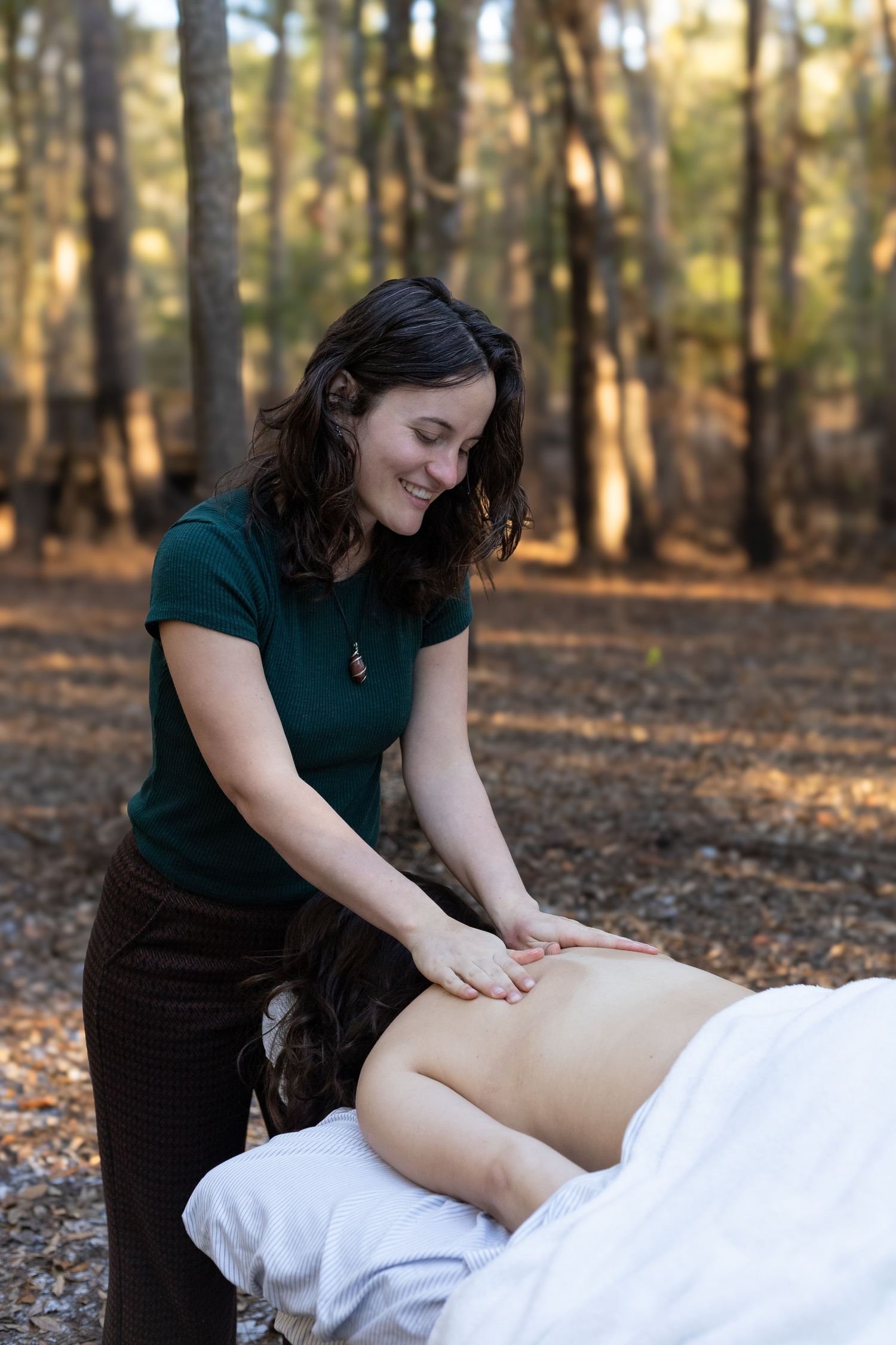 A smiling female massage therapist performs an outdoor back massage in a serene forest setting.