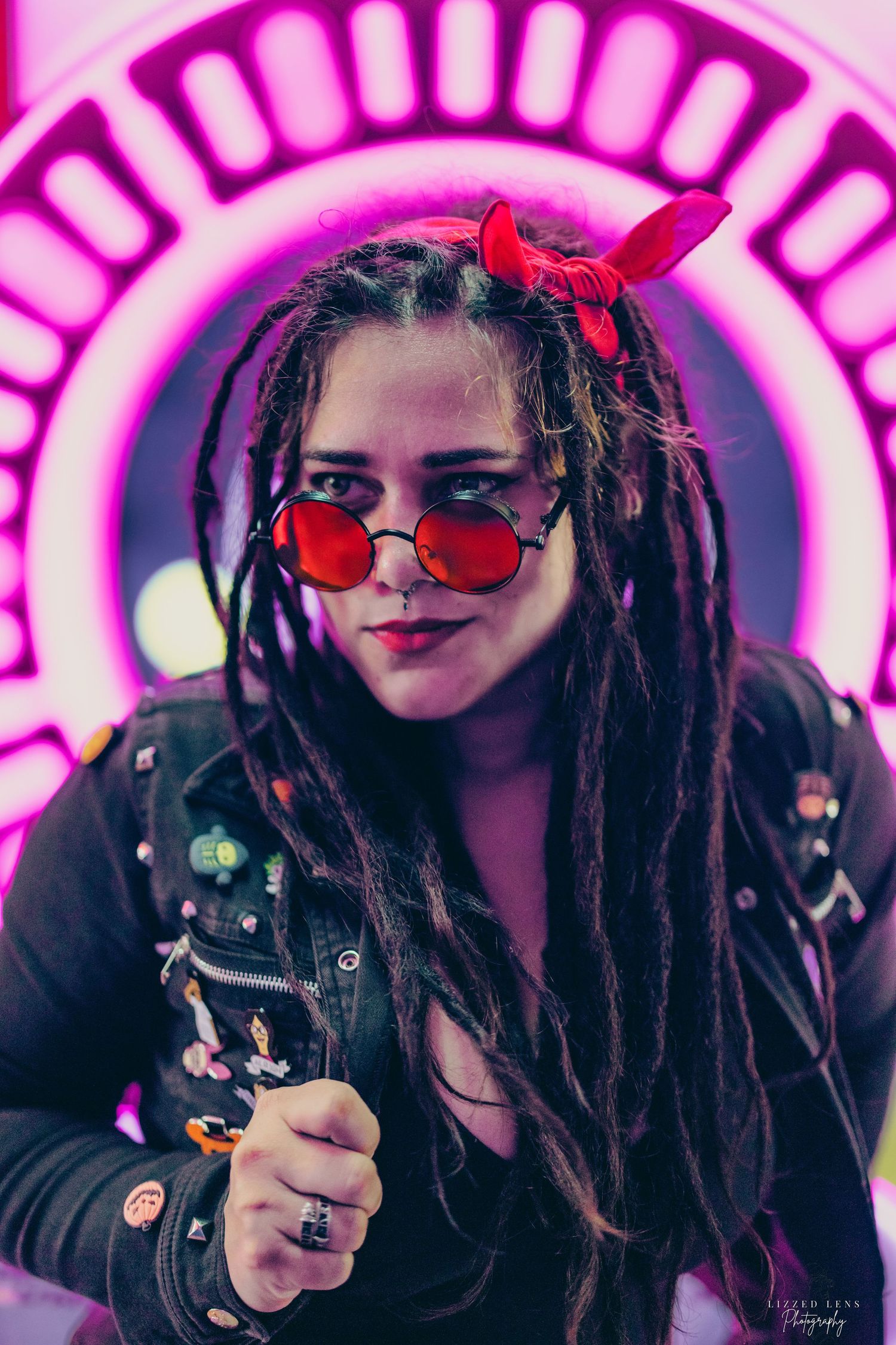 A woman with dreadlocks and red round glasses poses before a glowing pink neon circle.