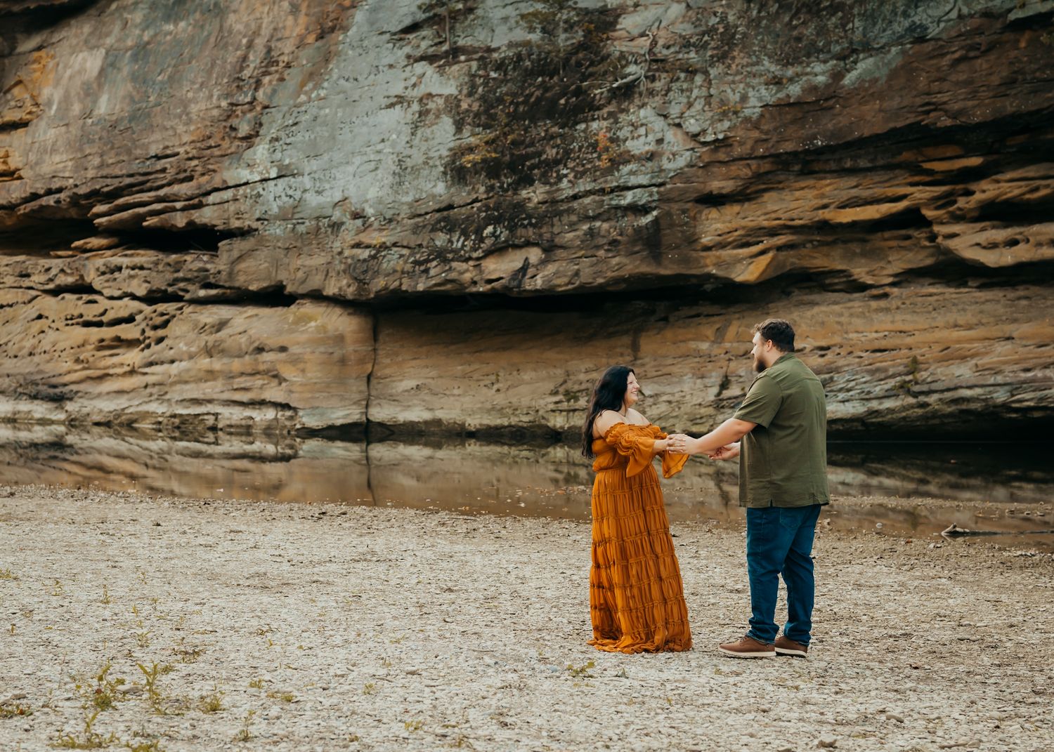 Couple stands, holding hands, making eye contact, in a riverbed, at turkey run state park, with a rock formation.