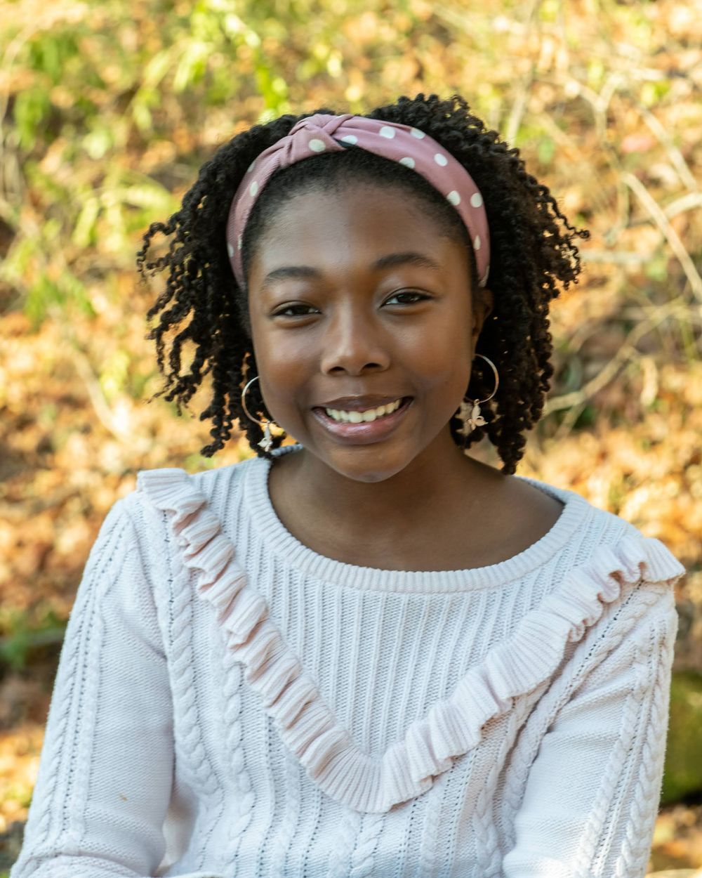 Portrait of young person in white sweater and pink headband against autumn background.