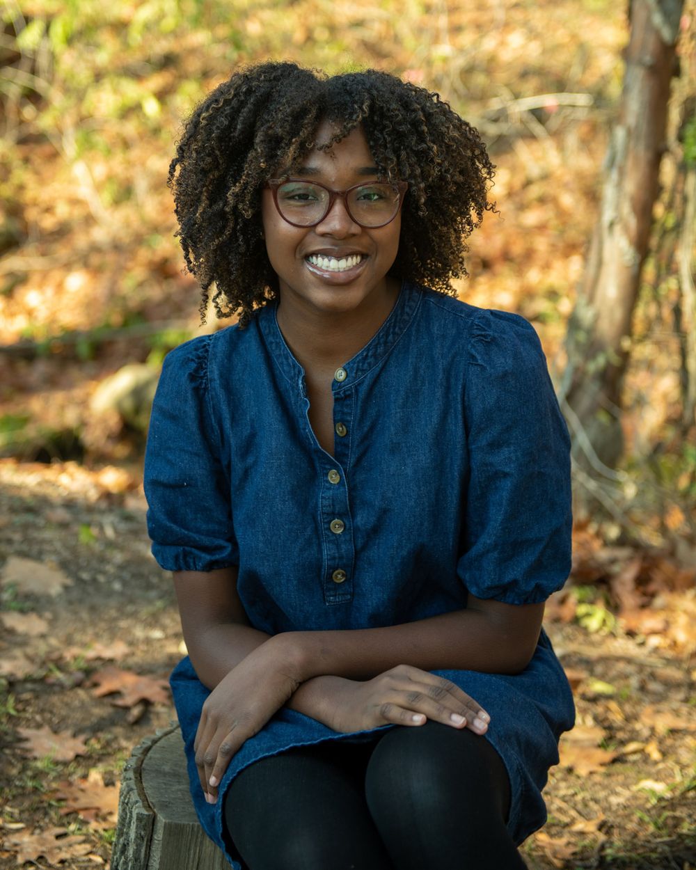Casual portrait sitting outdoors wearing denim top against autumn foliage.