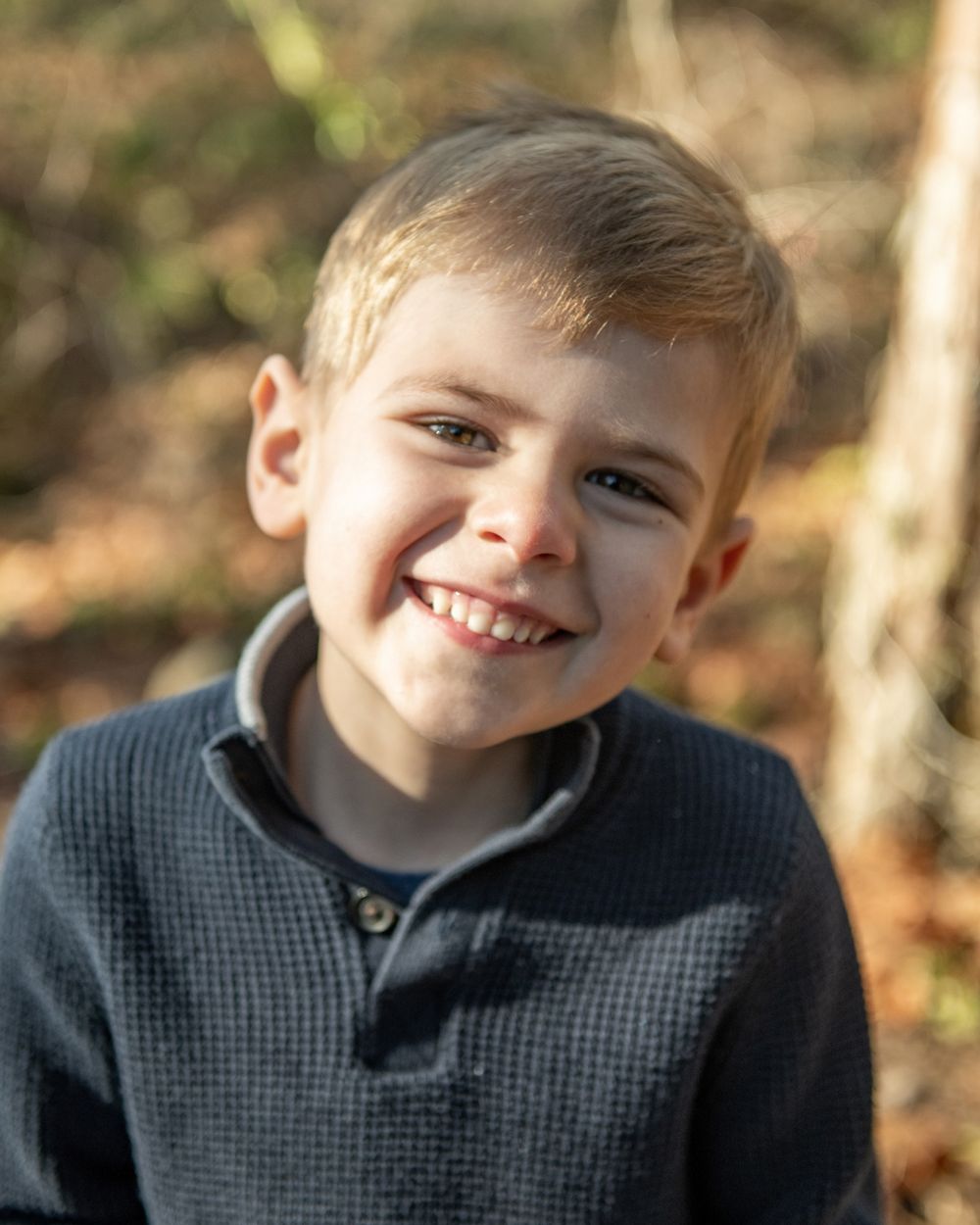 Young child in dark pullover smiles brightly against blurred autumn background.