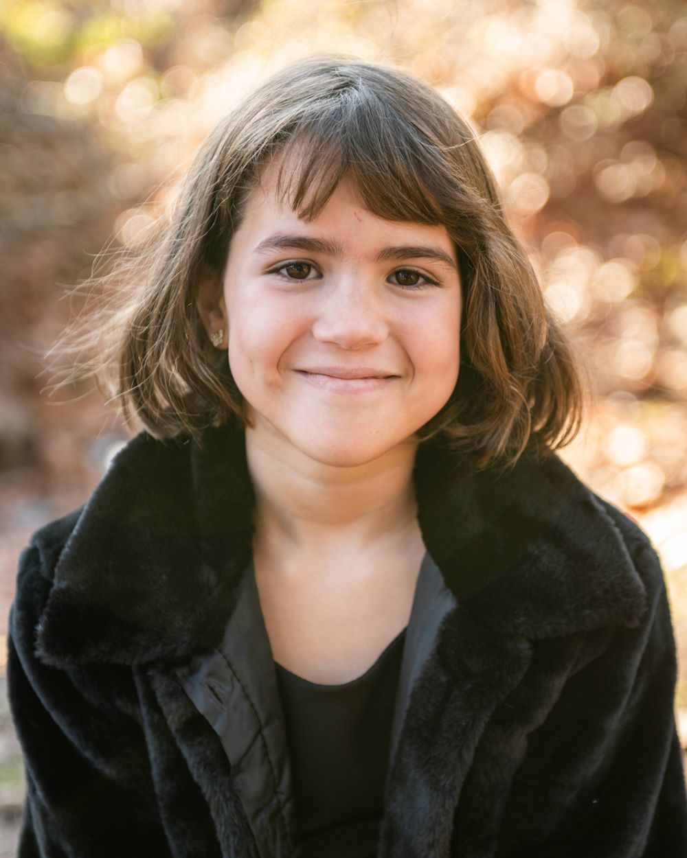 Child in black coat smiles warmly against soft golden bokeh background.