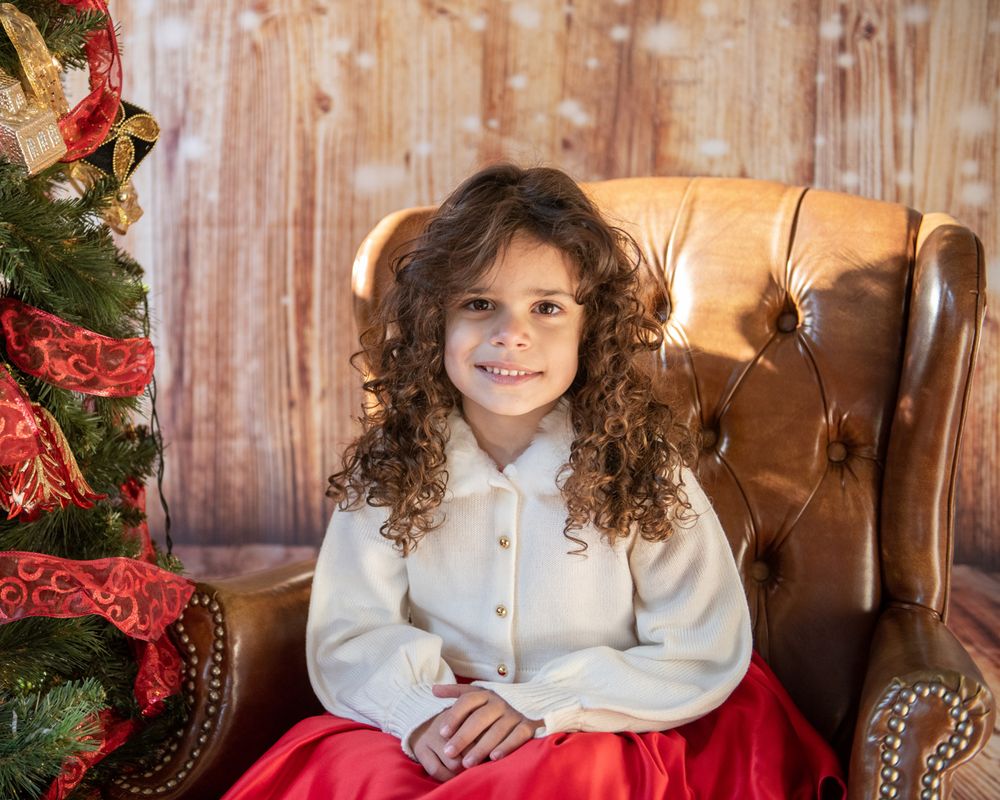 Child in white blouse and red skirt sitting near Christmas tree and leather chair.