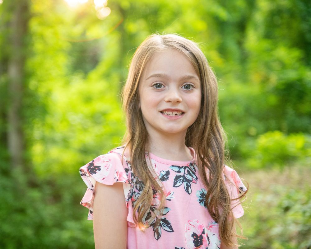 Child in pink floral dress smiles brightly against blurred green forest background in outdoor portrait.