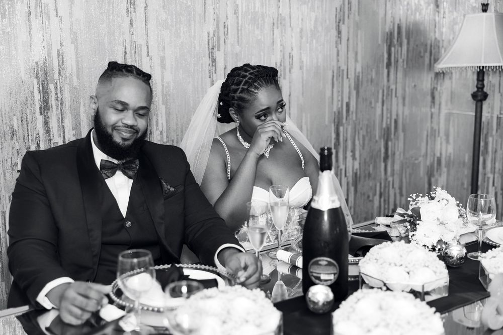 A black and white photo of a wedding couple dining at their reception with wine and flowers on the table.