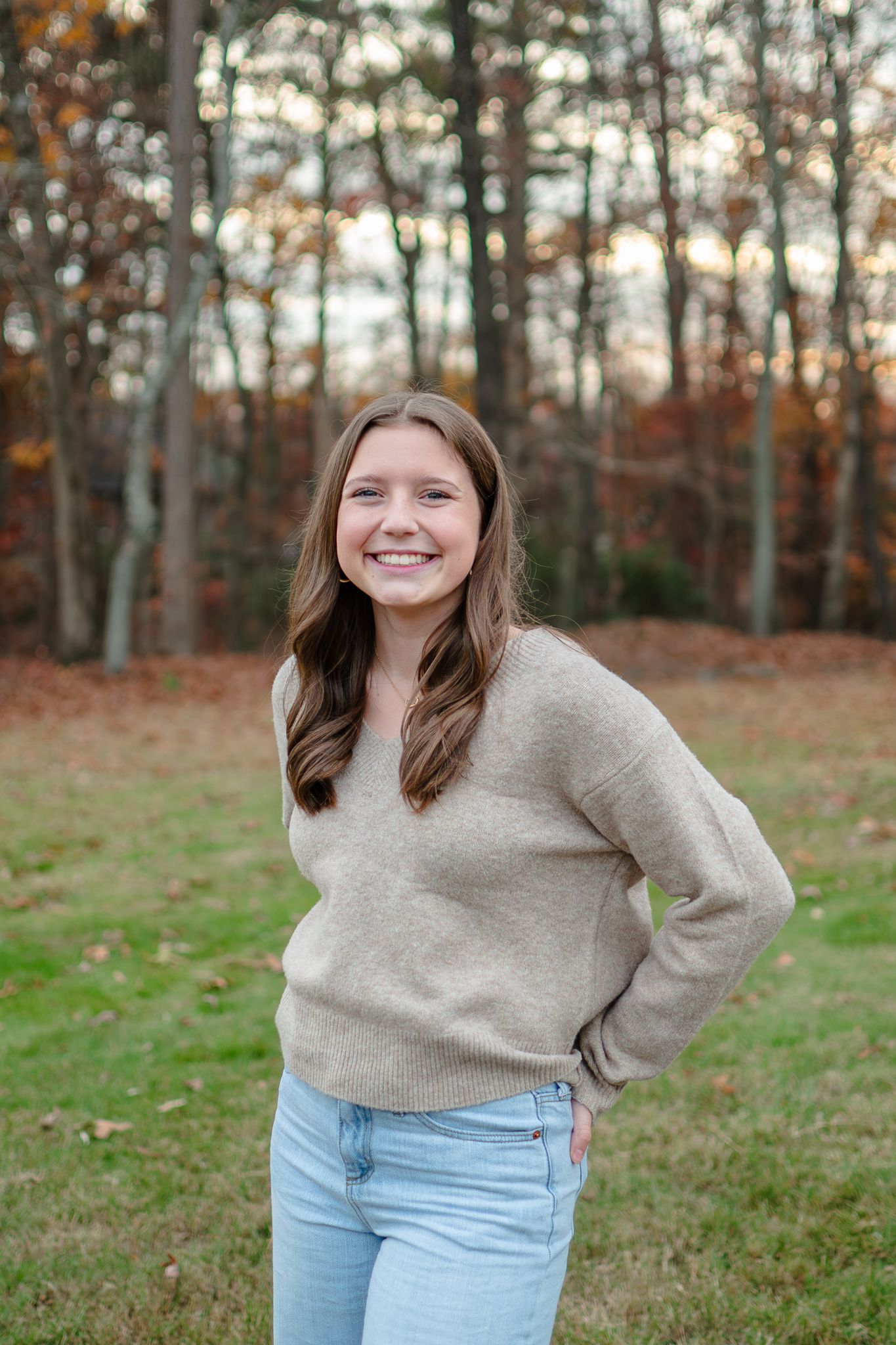 Casual autumn portrait with beige sweater and jeans standing in front of fall foliage and trees.