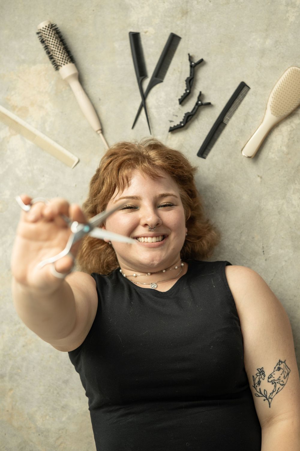 Scissors and hairstyling tools arranged around a person in black clothing against a textured wall.