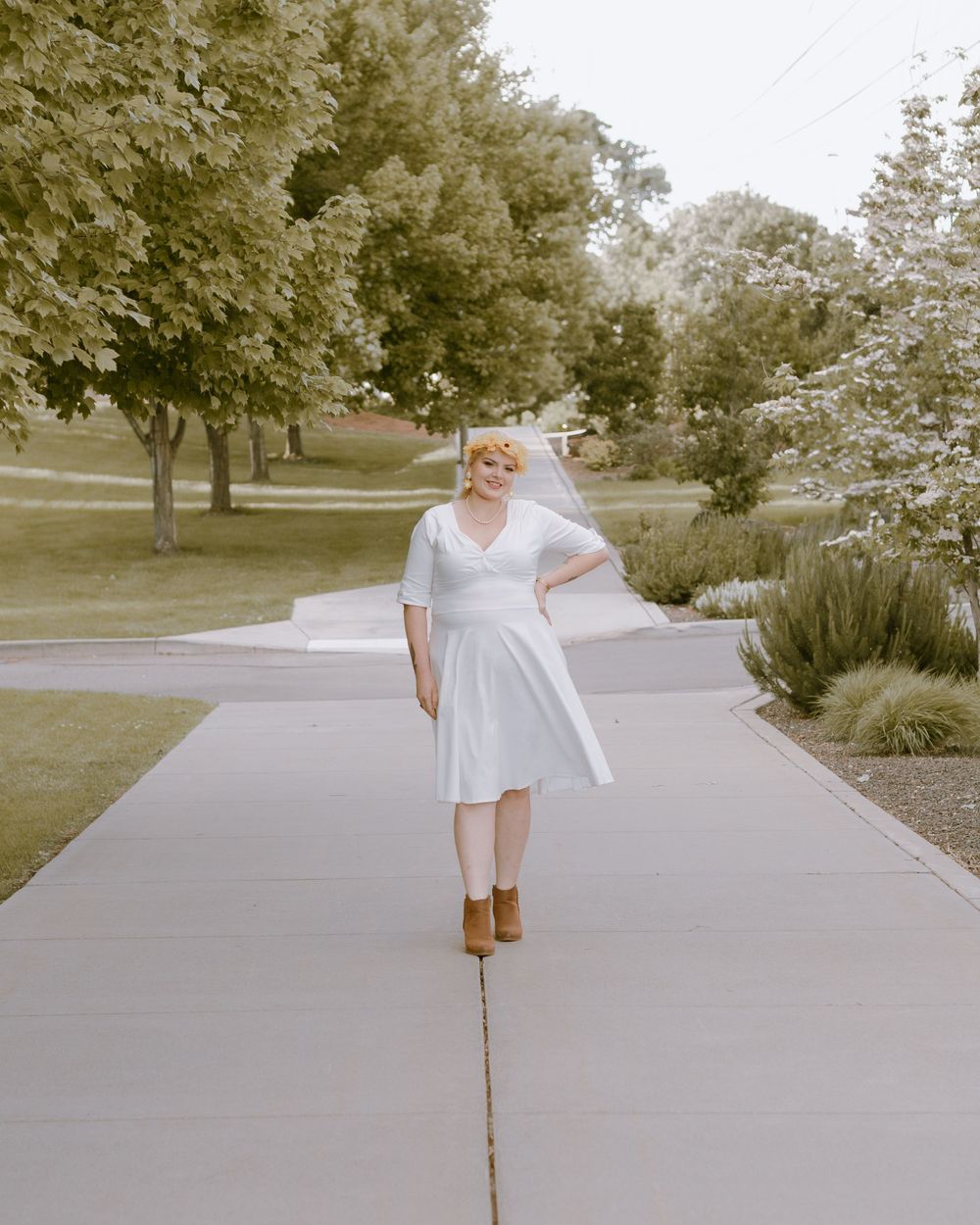 Woman in yellow flower crown, white dress, and brown booties posed on a tree-lined sidewalk.