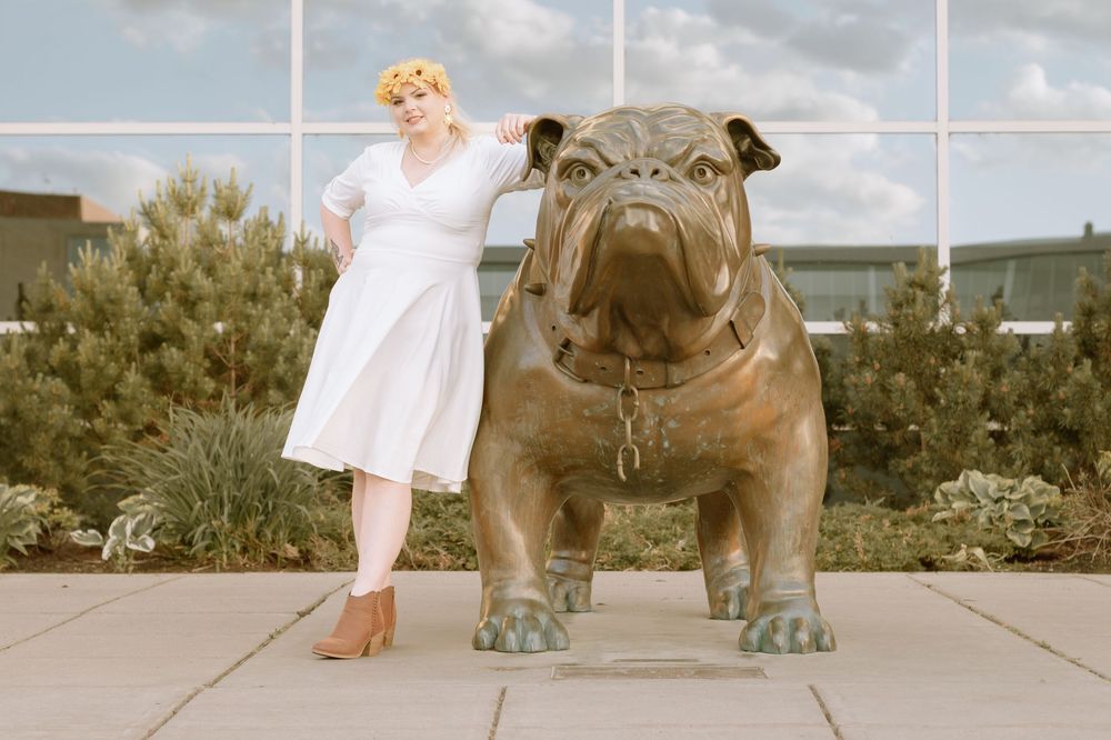 Woman in white dress wearing yellow flower crown posing next to a pug statue.
