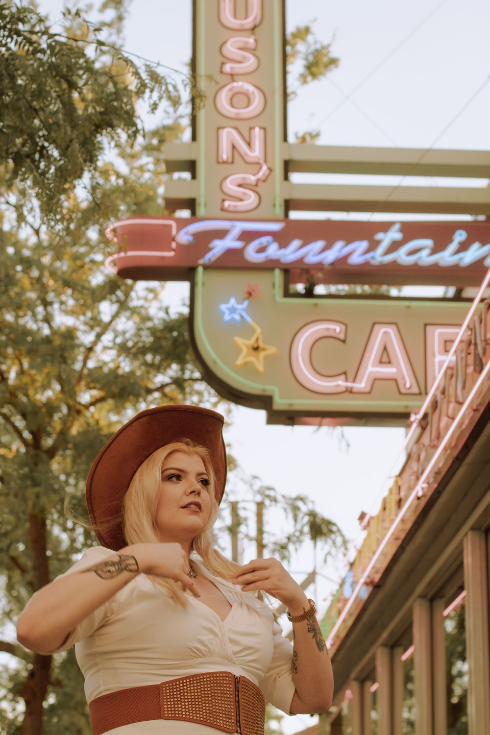 Blonde woman in casual western dress and a cowgirl hat fixing hair in front of Ferguson's Fountain Cafe.