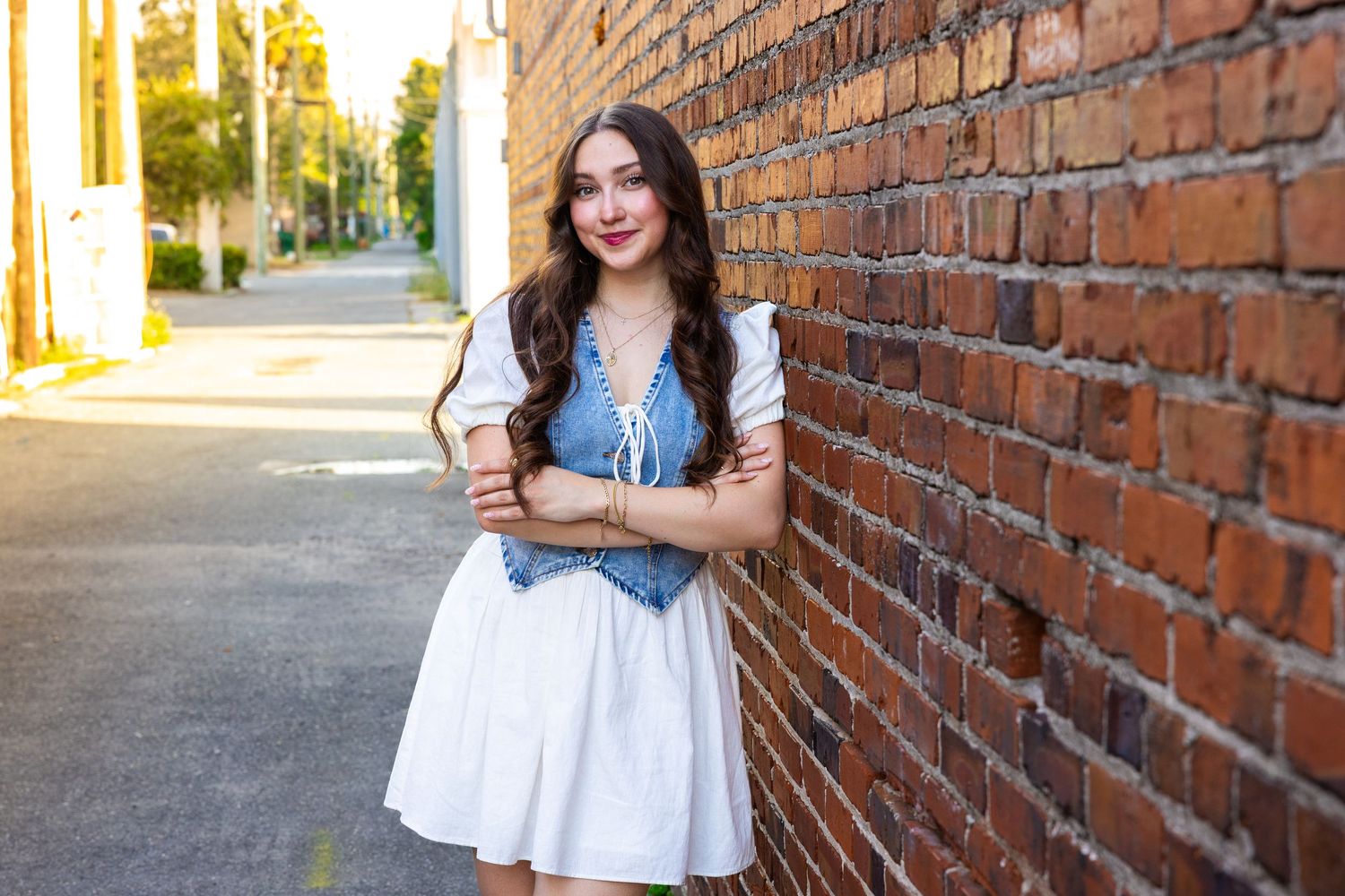 Woman in white dress and denim jacket poses against rustic brick wall backdrop in outdoor photoshoot.
