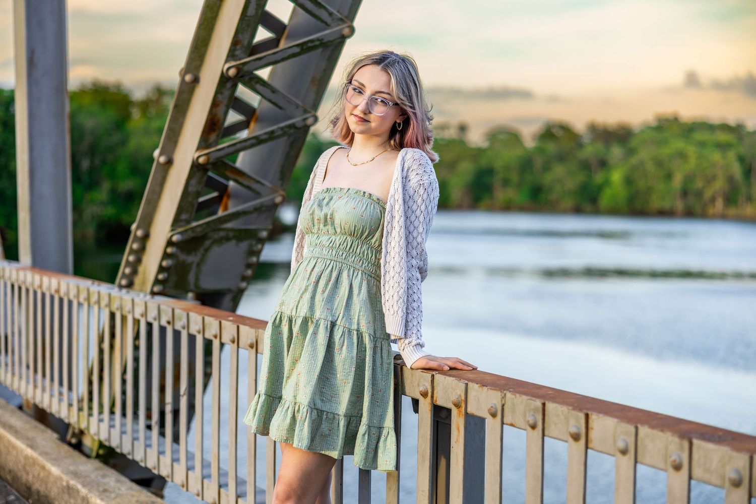 Woman in sage green dress poses outdoors on wooden boardwalk by scenic waterfront during golden hour.