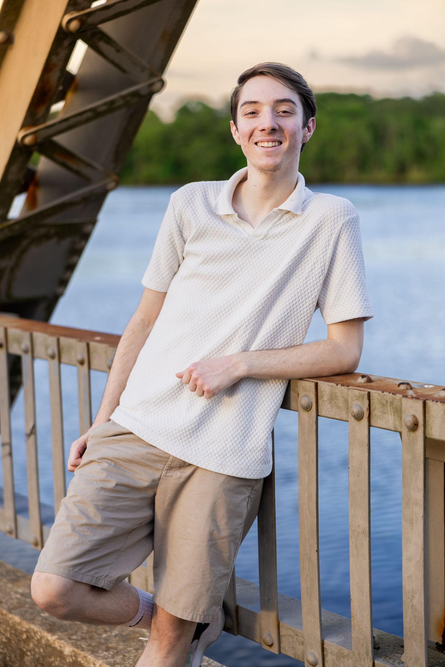 Person in beige shorts and white shirt poses casually against wooden railing by peaceful lakeside.