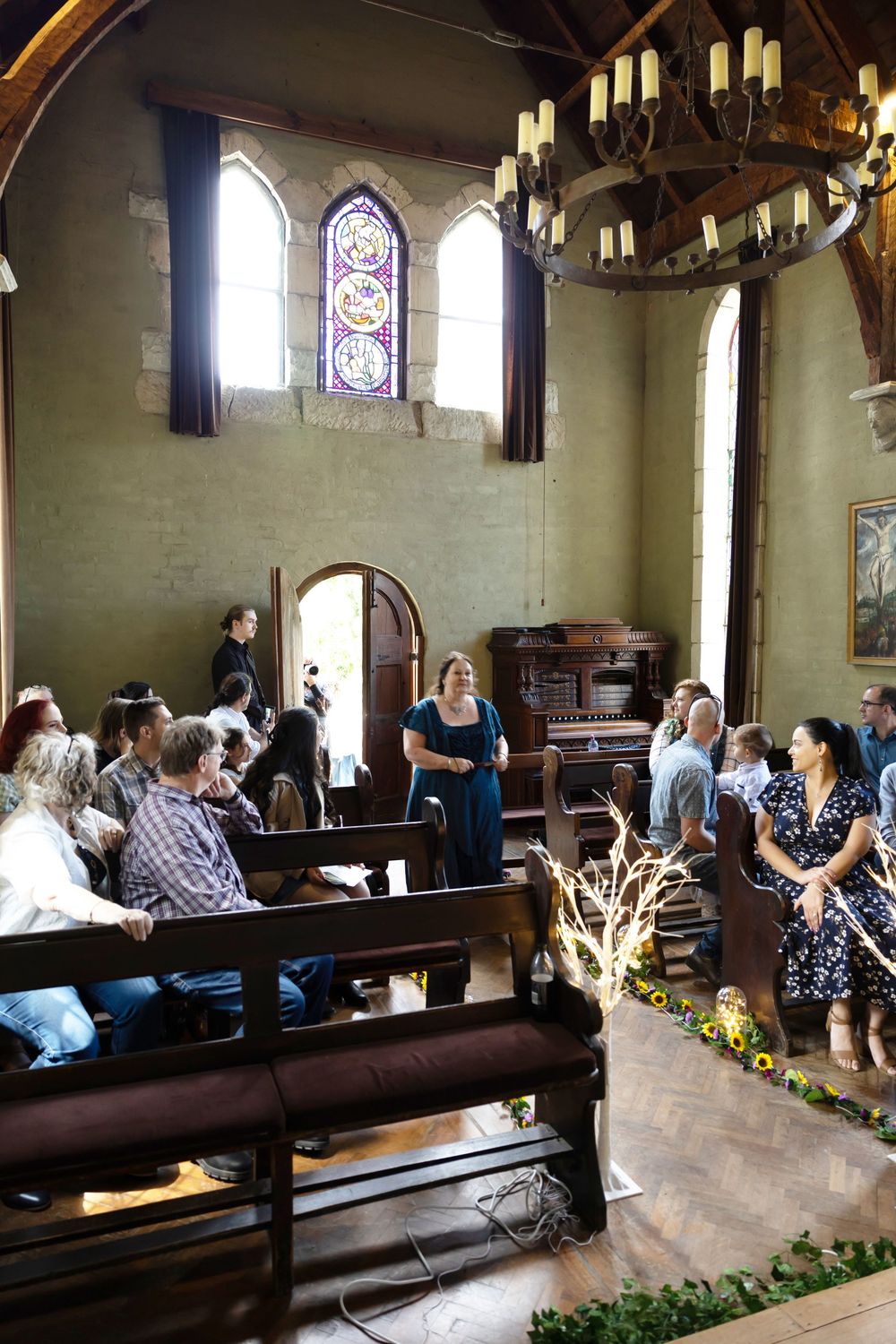 People gather inside a historic church interior with stained glass windows, wooden pews, and a rustic chandelier.