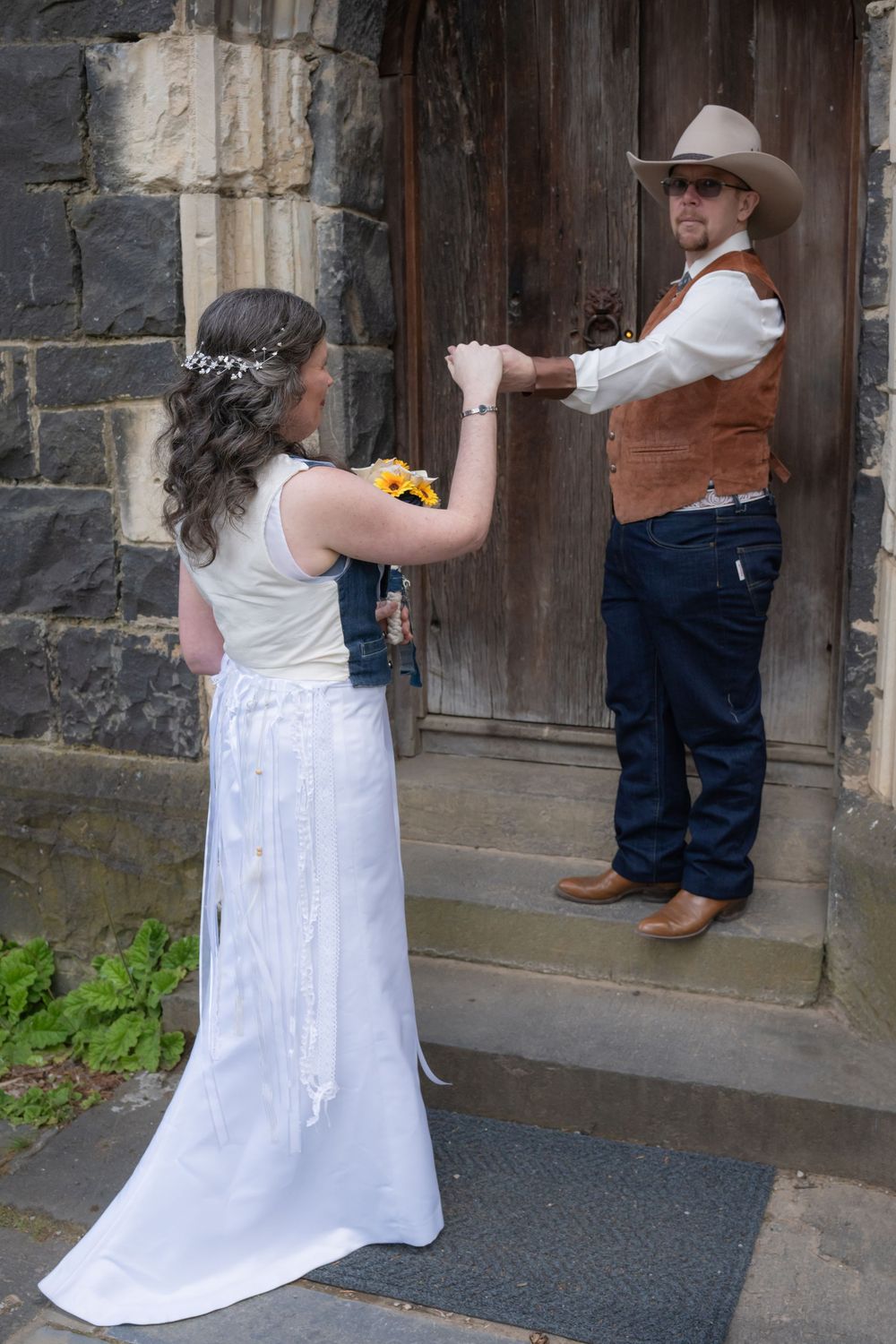 A couple in western wedding attire stands at an old wooden door entrance to a stone church.