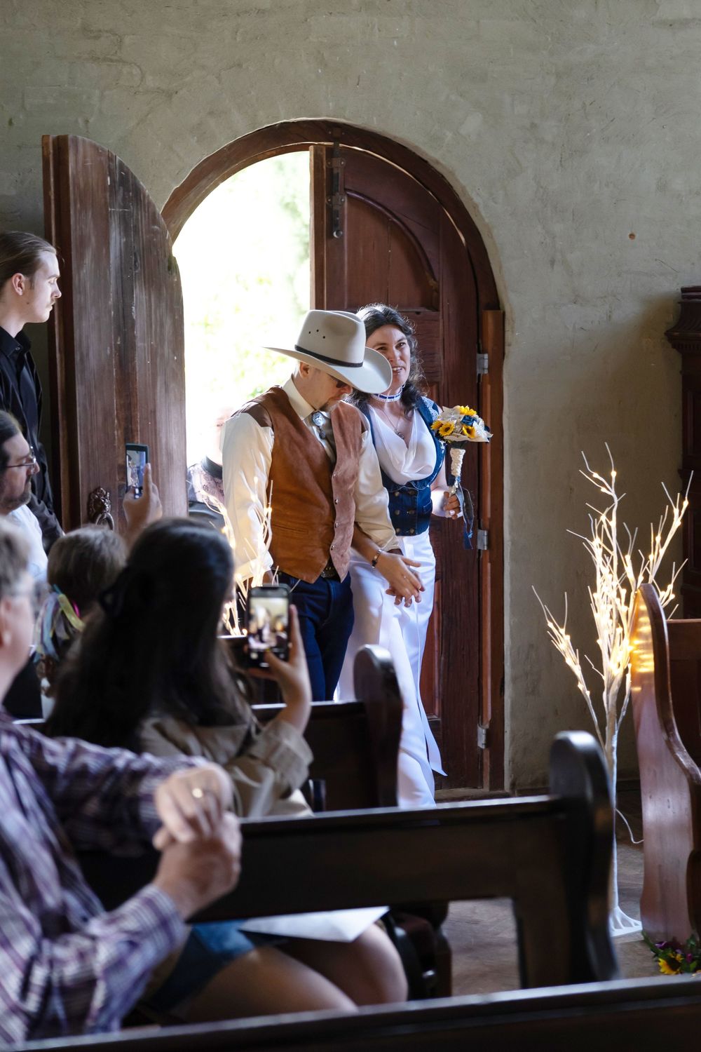 Guests rise from wooden church pews during an indoor wedding ceremony at a rustic venue with stone walls.