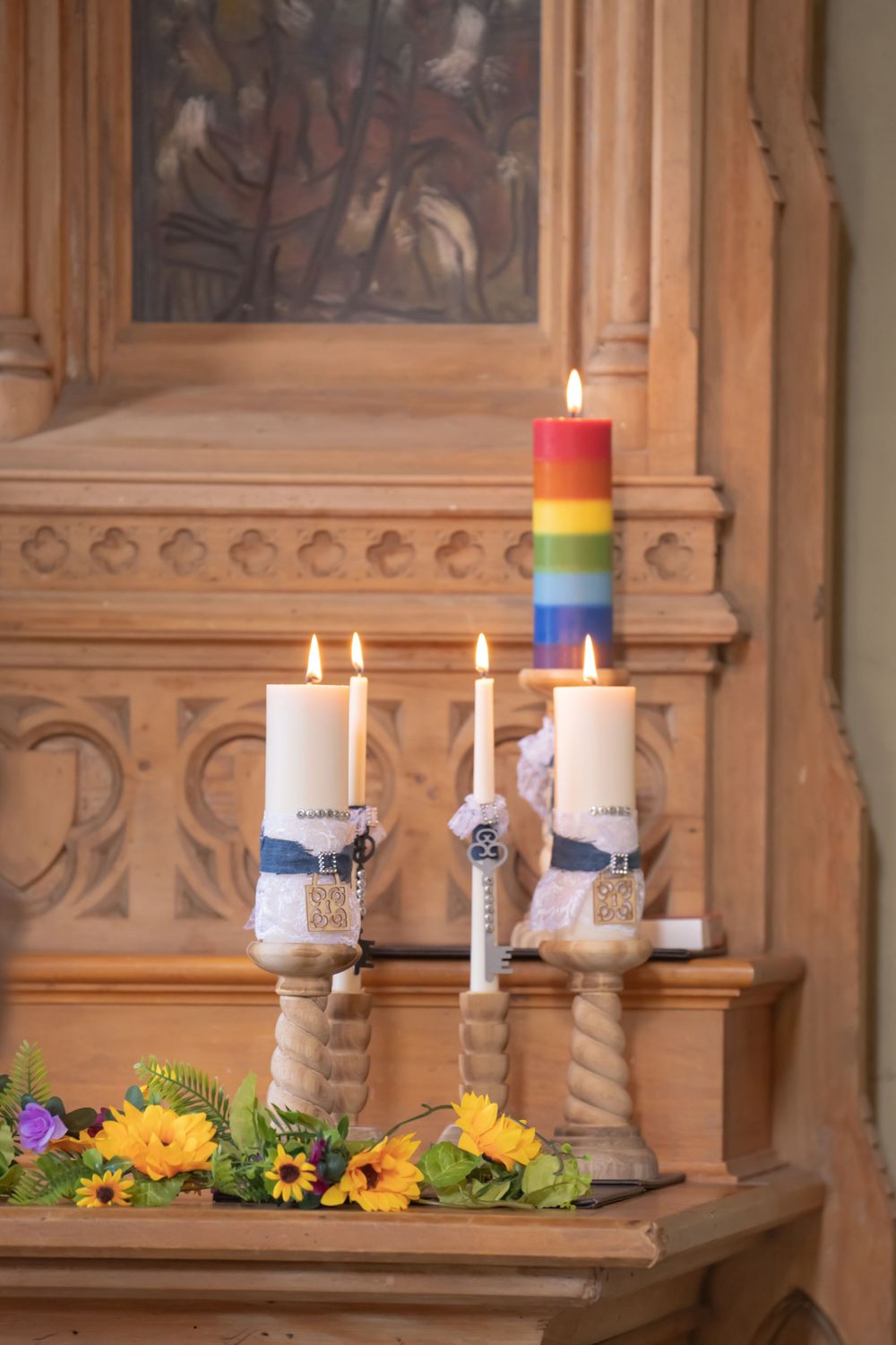 Rainbow pride candles and yellow flowers arranged on a wooden church altar with gothic architectural details.