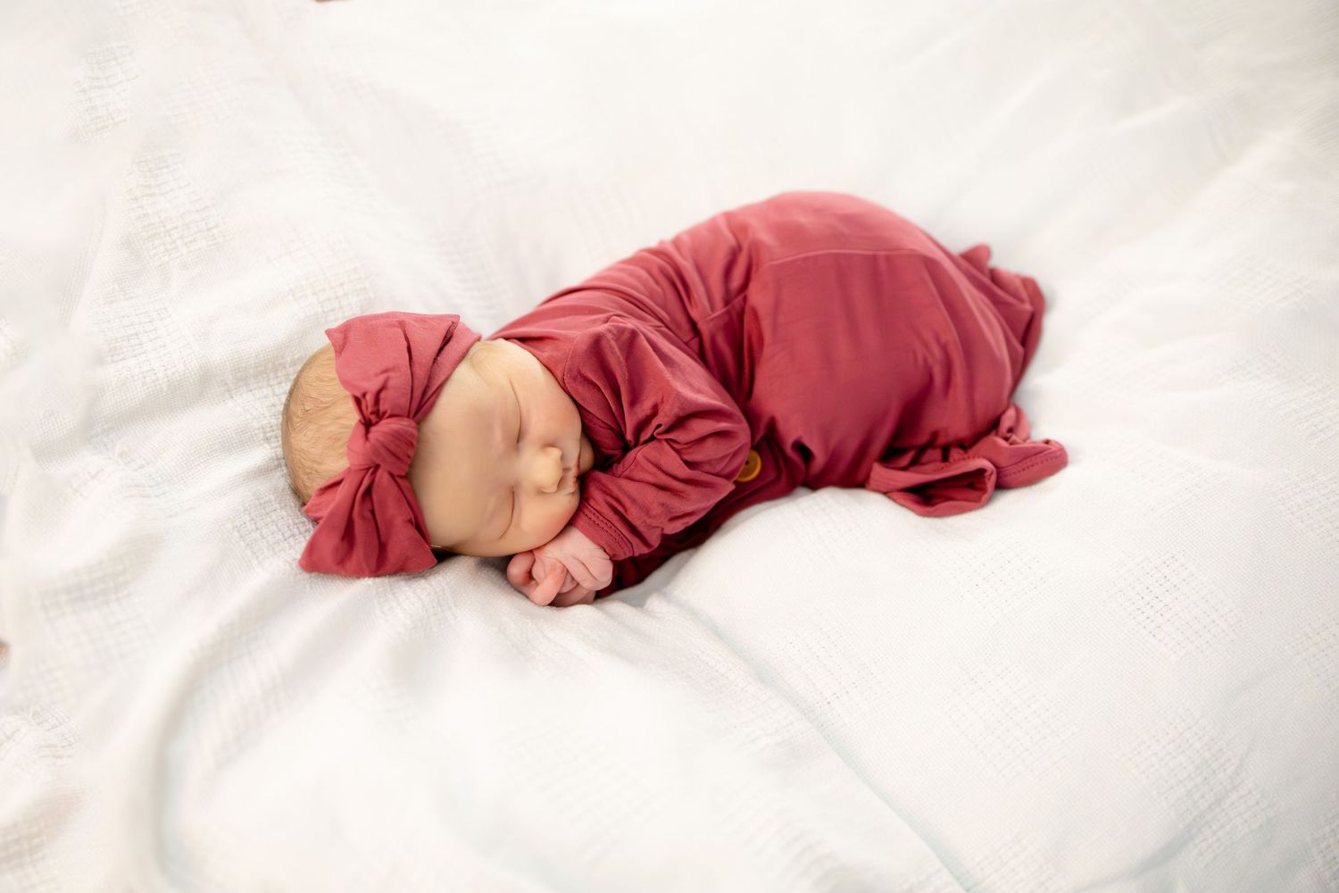 Newborn baby sleeping peacefully in a red outfit and matching bow headband on a white textured blanket.