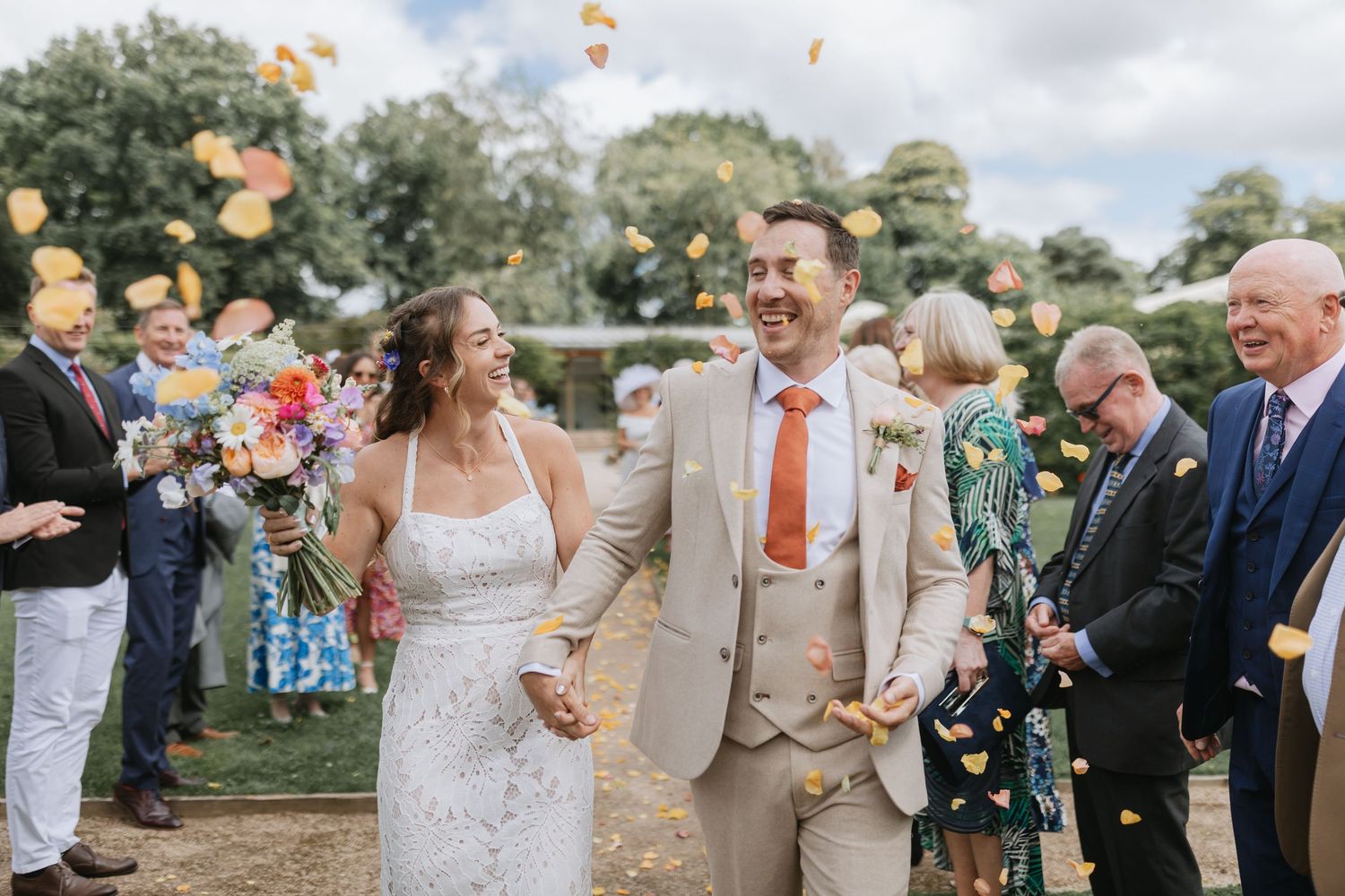 Wedding celebration with guests tossing orange flower petals in a joyful moment by a lake at an outdoor ceremony.