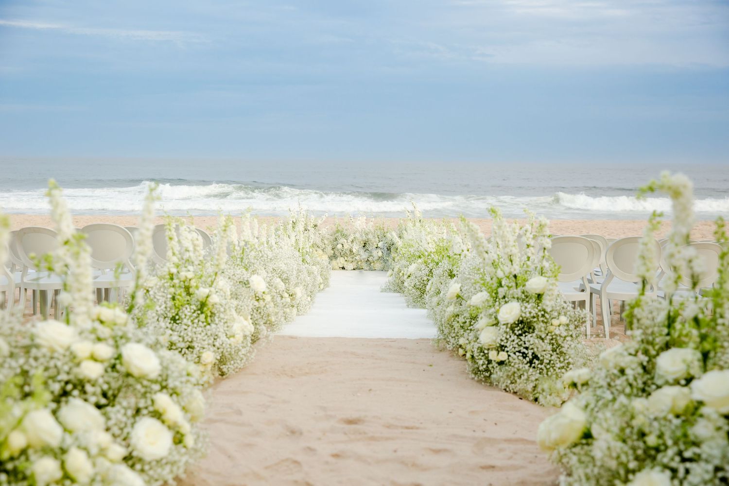 Beach wedding ceremony setup with white flowers lining the aisle against ocean backdrop.