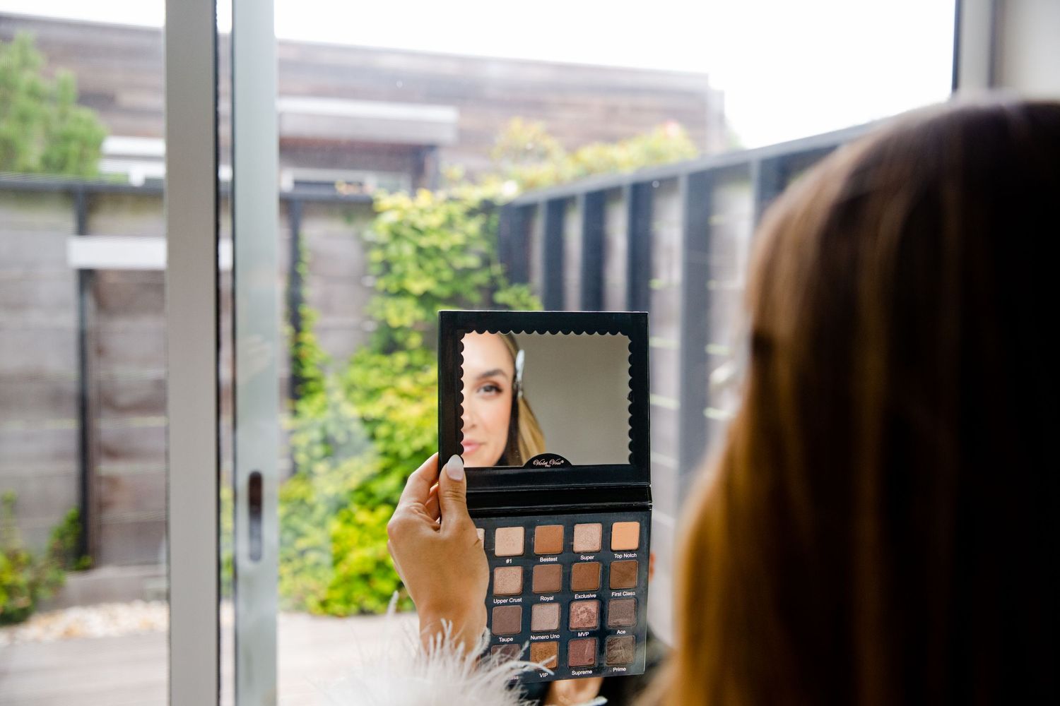 Person applying makeup using a compact mirror near a window with garden view.