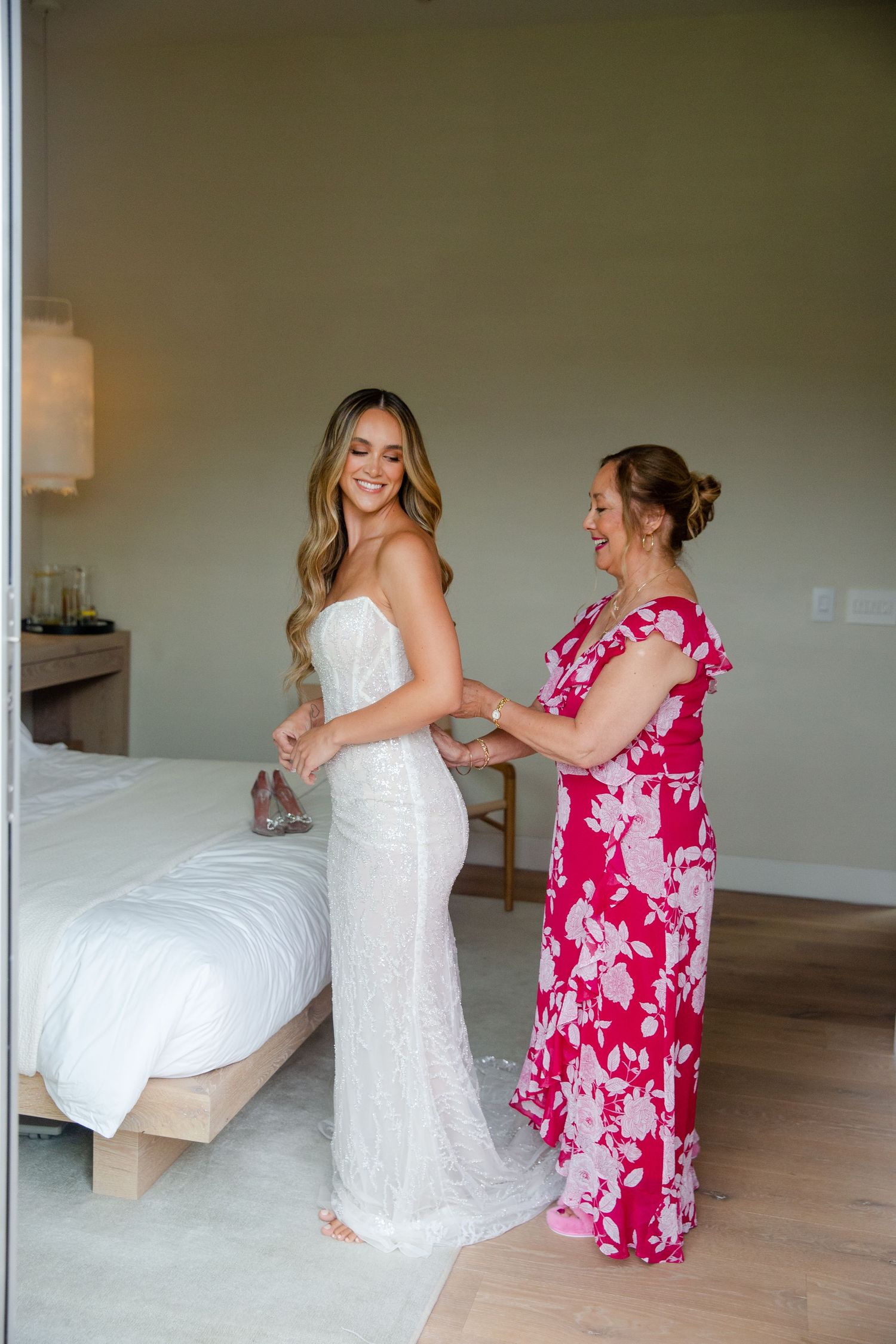 A bride in a fitted white lace gown gets help from someone wearing a vibrant pink floral dress in a minimalist room.