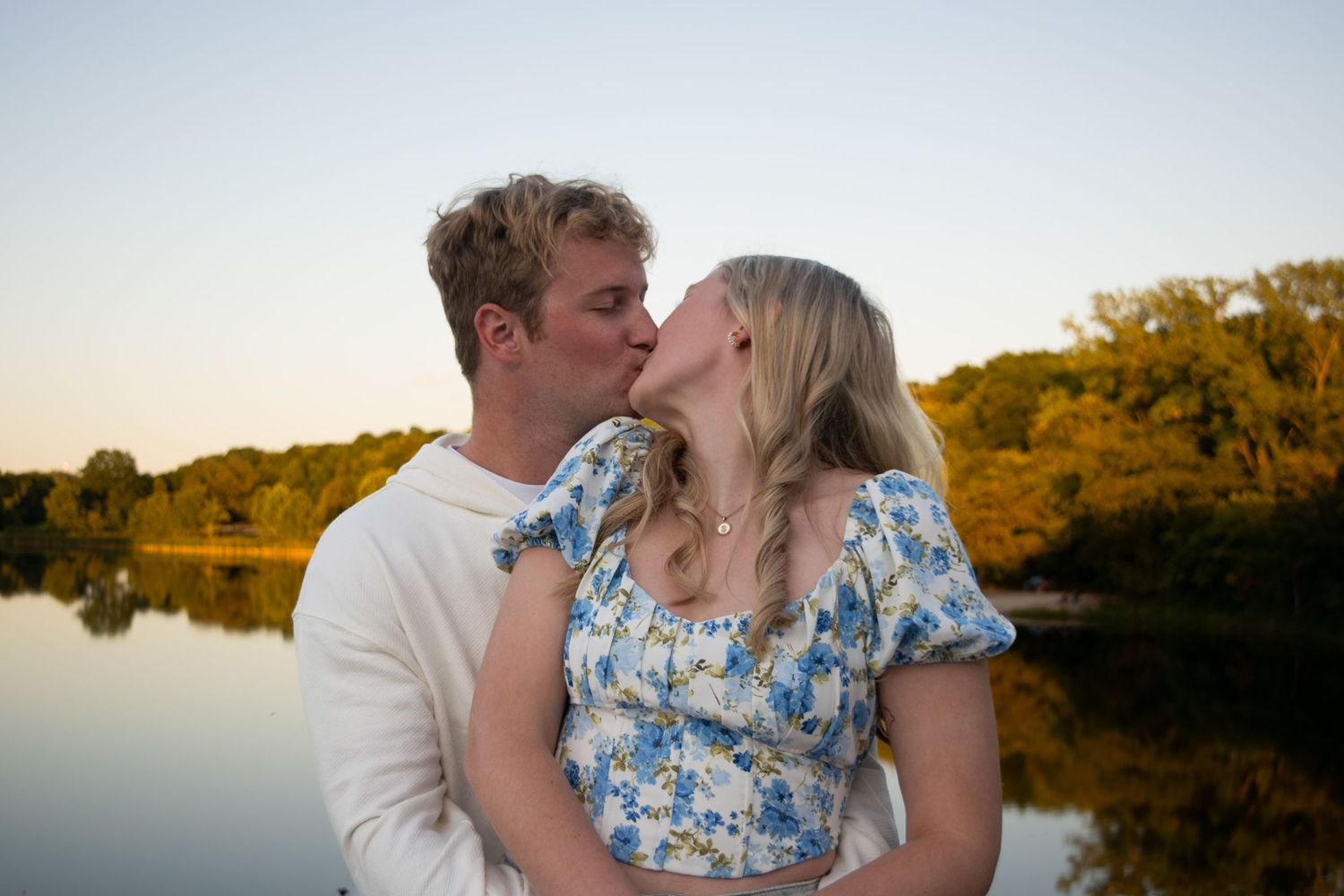 Woman standing in front of a man with her head turned back to kiss him with a lake in the background at sunset